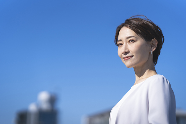 A middle-aged Japanese woman wearing a suit against a sunny blue sky background