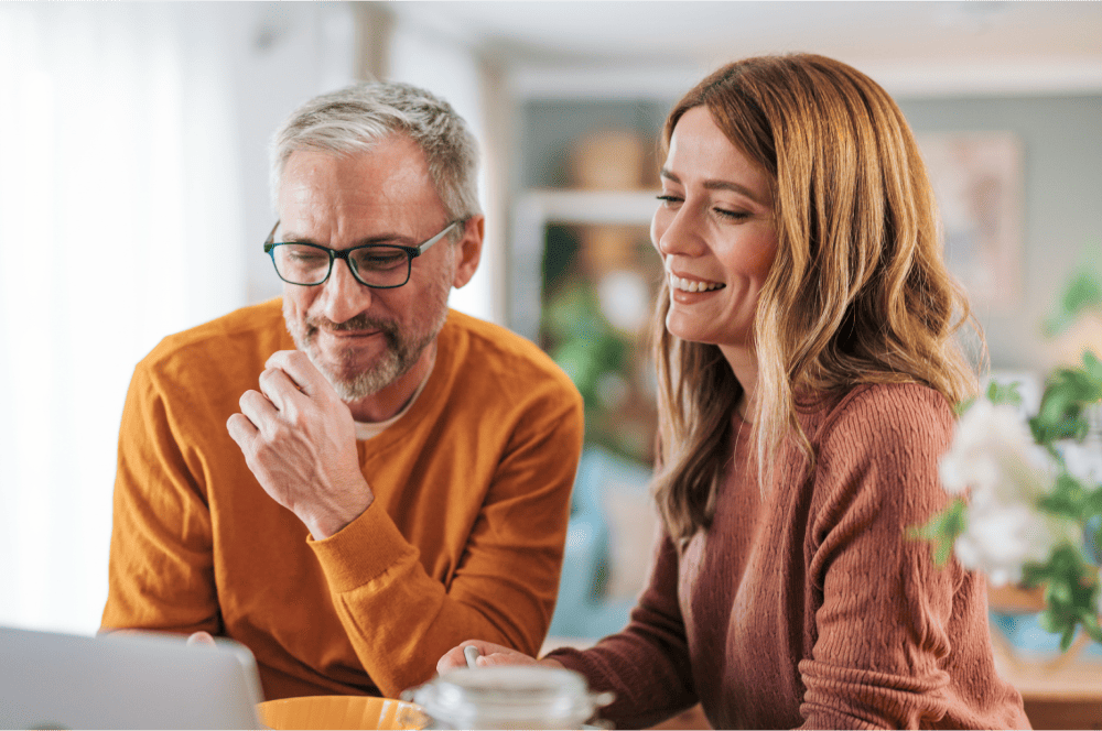Middle-aged couple, sat at a table and smiling at a laptop 