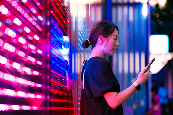 A woman looking at a smartphone in front of red neon lights, Getty