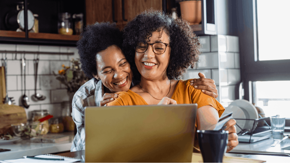 Two women in a kitchen looking at a laptop and smiling